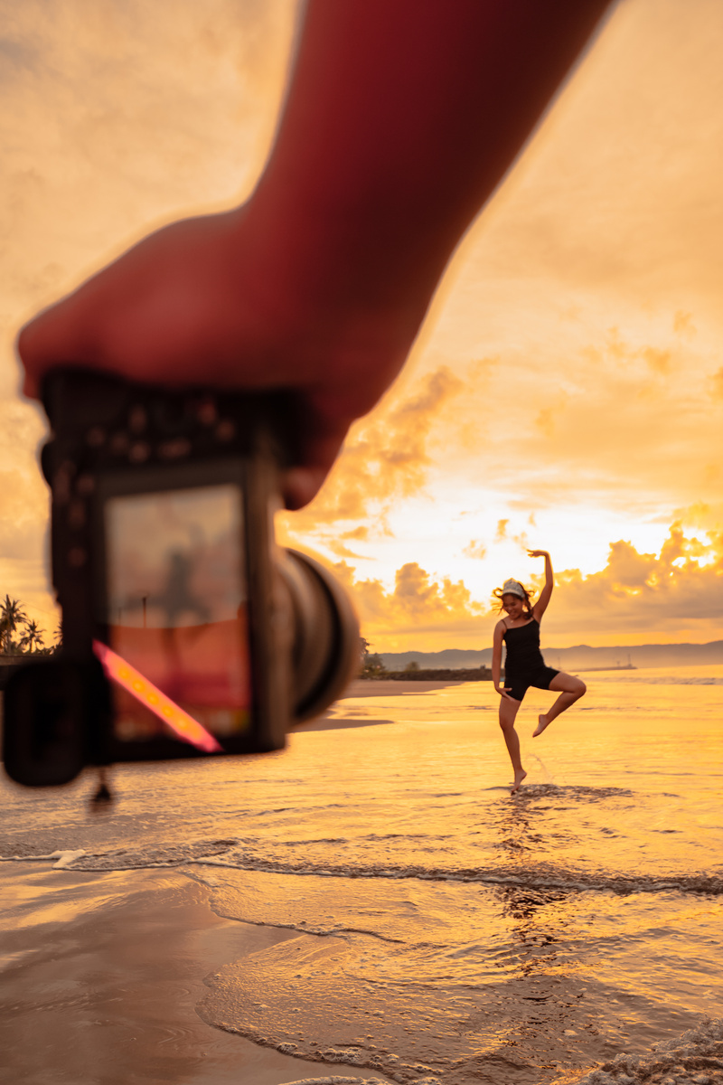 A Camera with hands photographs a Balinese woman doing a gymnastic movement on a black shirt near the beach